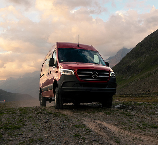 A Mercedes-Benz Camper Van parked on a steep hill with a person sitting next to it fixing their bike. 