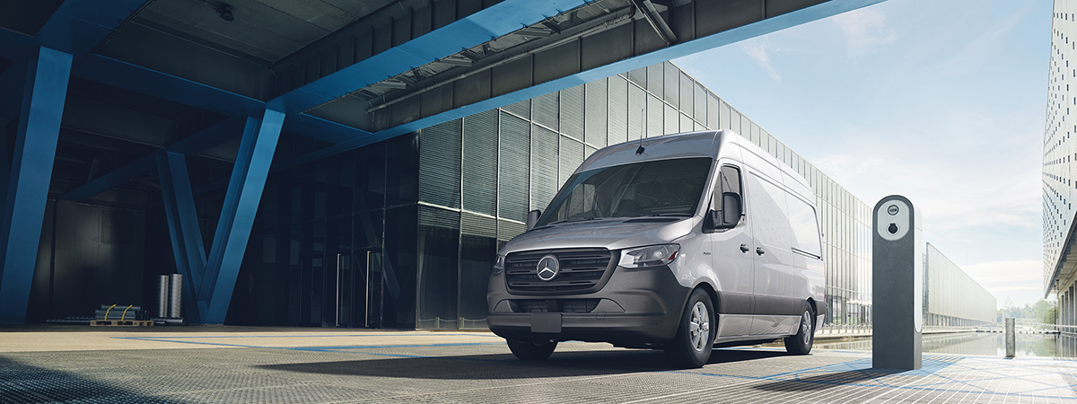 A wide shot of a silver Mercedes-Benz eSprinter Cargo van parked in front of a grey industrial building. 